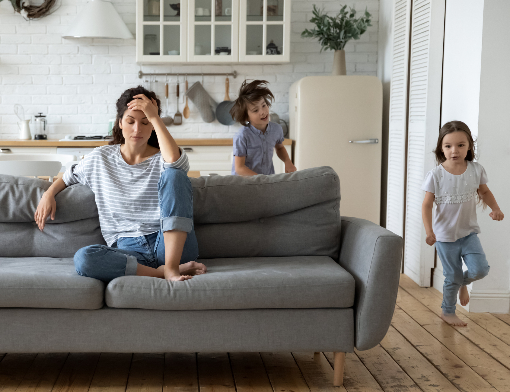 Mulher sentada no sofá com as mãos na testa enquanto os filhos brincam em casa